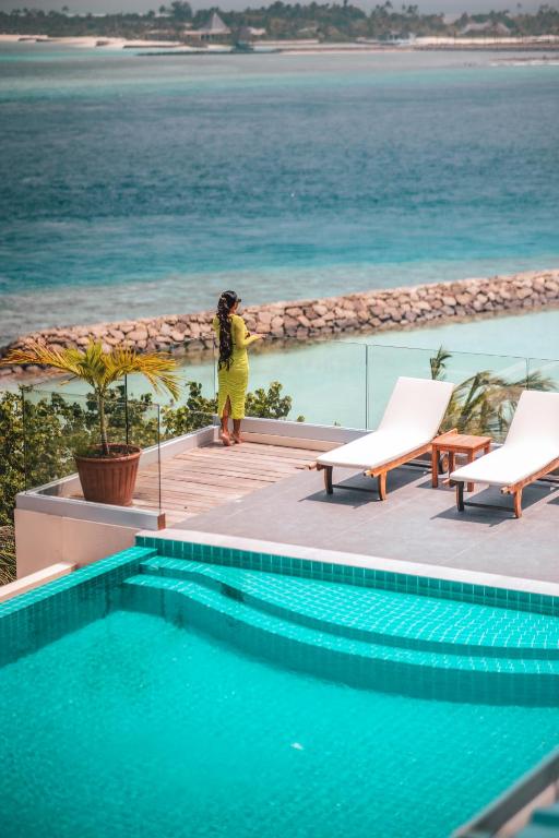 Woman in a yellow dress stands on a resort deck, looking out over turquoise ocean, with a pool and white lounge chairs nearby.