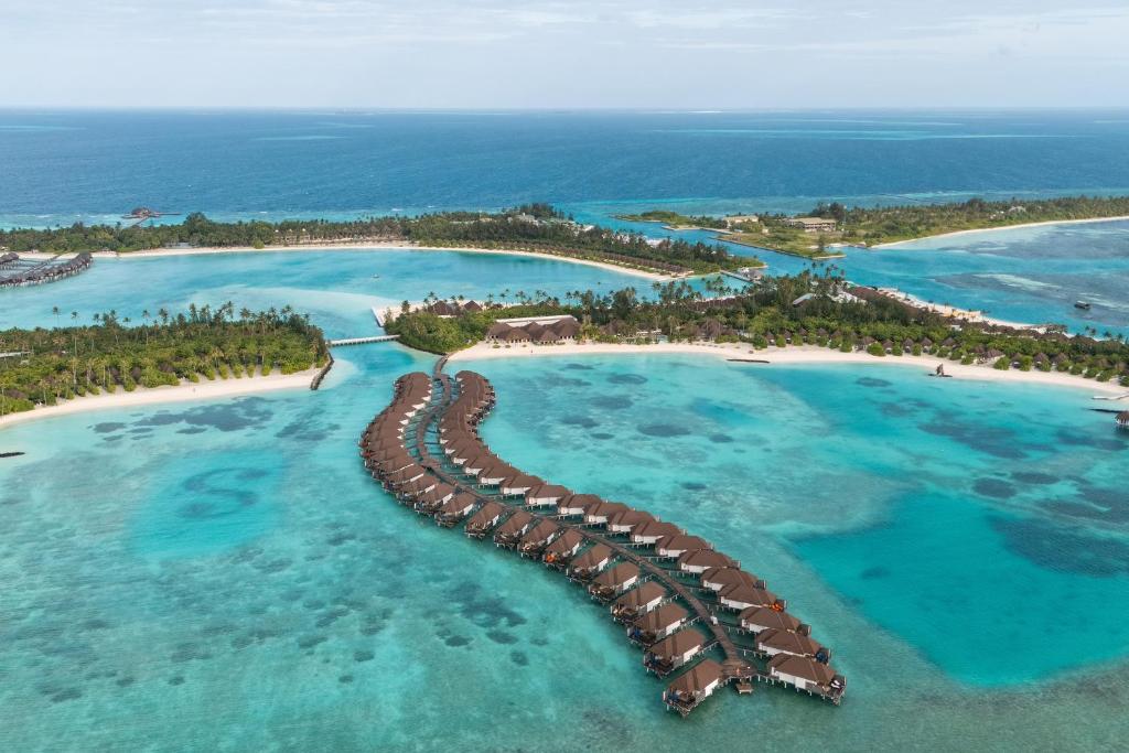 Overwater bungalows curve along a turquoise lagoon toward a sandy beach at a tropical resort island.",