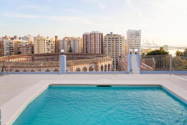 Rooftop swimming pool overlooking a city skyline with mid-rise buildings and a distant harbor on the right.