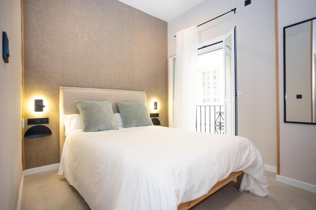 Hotel bedroom with a white duvet, two green pillows, and a grey textured accent wall; bedside lamps, open balcony door with iron railing, sheer white curtains, and a full-length mirror on the right.
