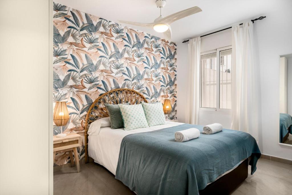 Bedroom with a palm-patterned accent wall, wicker headboard, and a double bed with a blue coverlet; two rolled towels on the bed.