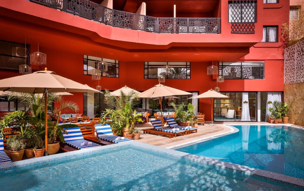 Poolside lounge area at a red hotel building, blue-striped chairs under beige umbrellas with potted plants. Temporary outdoor seating by the pool.