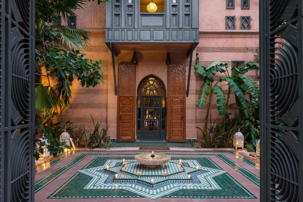 Moroccan-style courtyard with a geometric star-patterned tiled fountain, lush plants, and wooden arched doors framed by warm lantern light.
