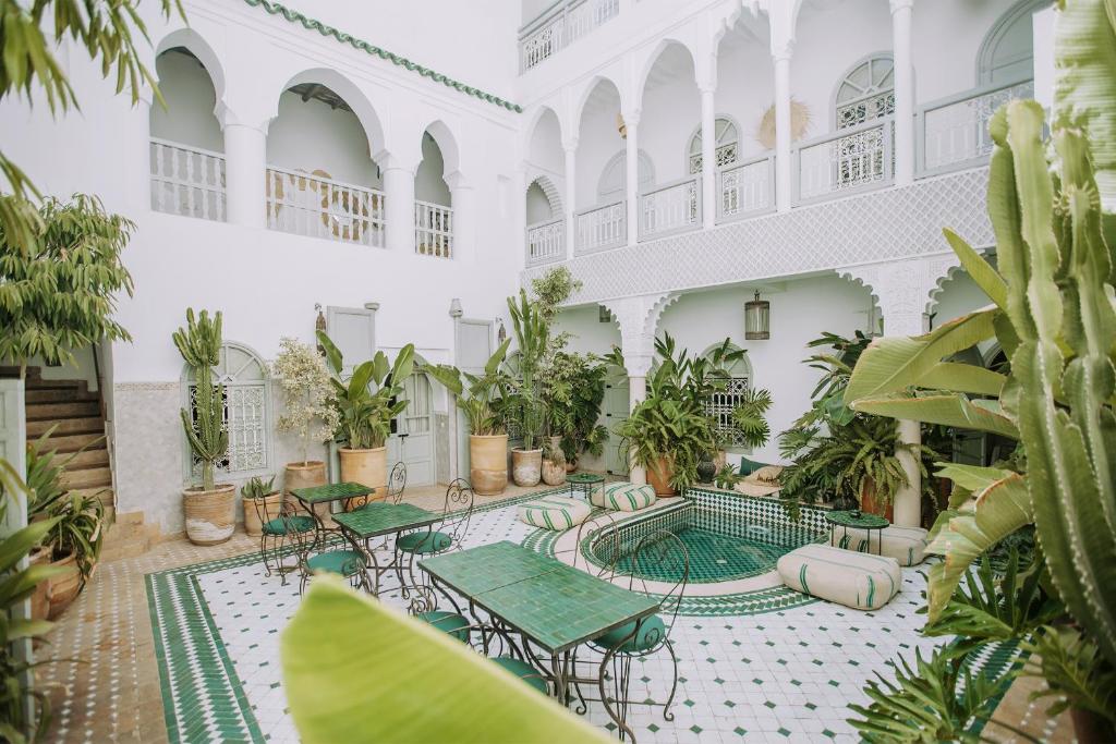 White-walled courtyard with arches, potted plants, and a small tiled pool surrounded by green tables and chairs.