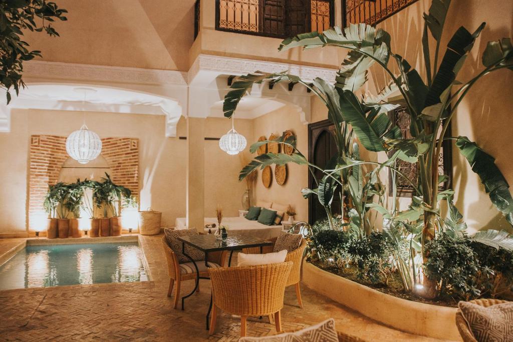 Indoor courtyard at night with a rectangular pool, wicker chairs around a glass table, and large tropical plants illuminated by warm lights.