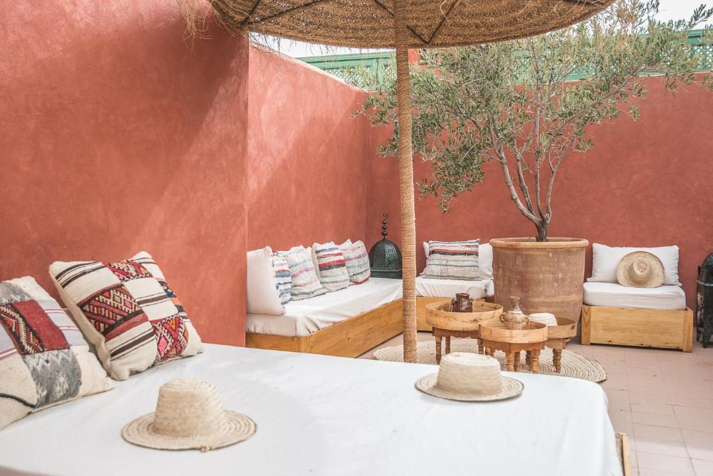 Outdoor courtyard seating with white cushions and colorful pillows against terracotta walls, a woven straw hat on a table, and a potted tree under a thatched umbrella.