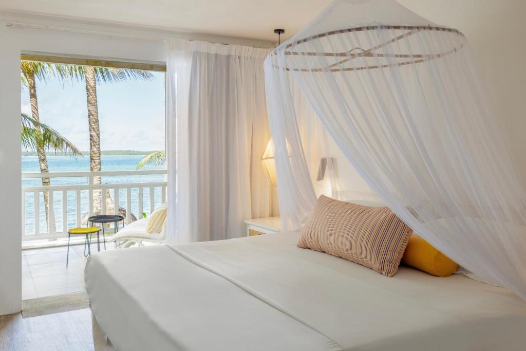 Bright hotel room with a white four-poster bed and sheer canopy, facing a balcony with an ocean view and palm trees outside.