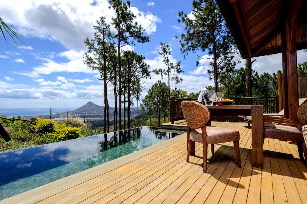 Outdoor wooden deck with a rectangular infinity pool, pine trees, and a distant mountain view; a dining table with wicker chairs sits to the right.