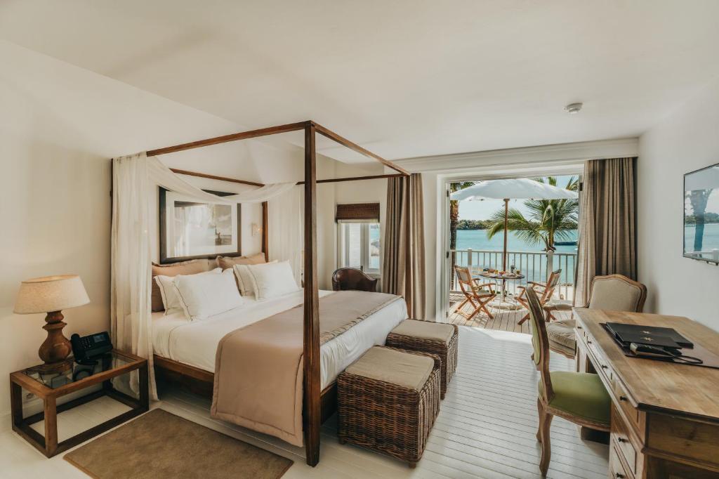 Luxurious hotel room with a dark wood four-poster bed, sheer drapes, and a balcony overlooking a tropical sea. Two wicker benches sit at the foot of the bed; a desk and chair are to the right. Outside, a table and chairs shaded by an umbrella.
