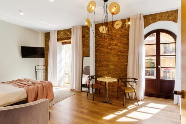 Bright hotel room with an exposed brick wall, bed with a peach throw, and a small round-table seating area by tall windows and curtains with a balcony door nearby
