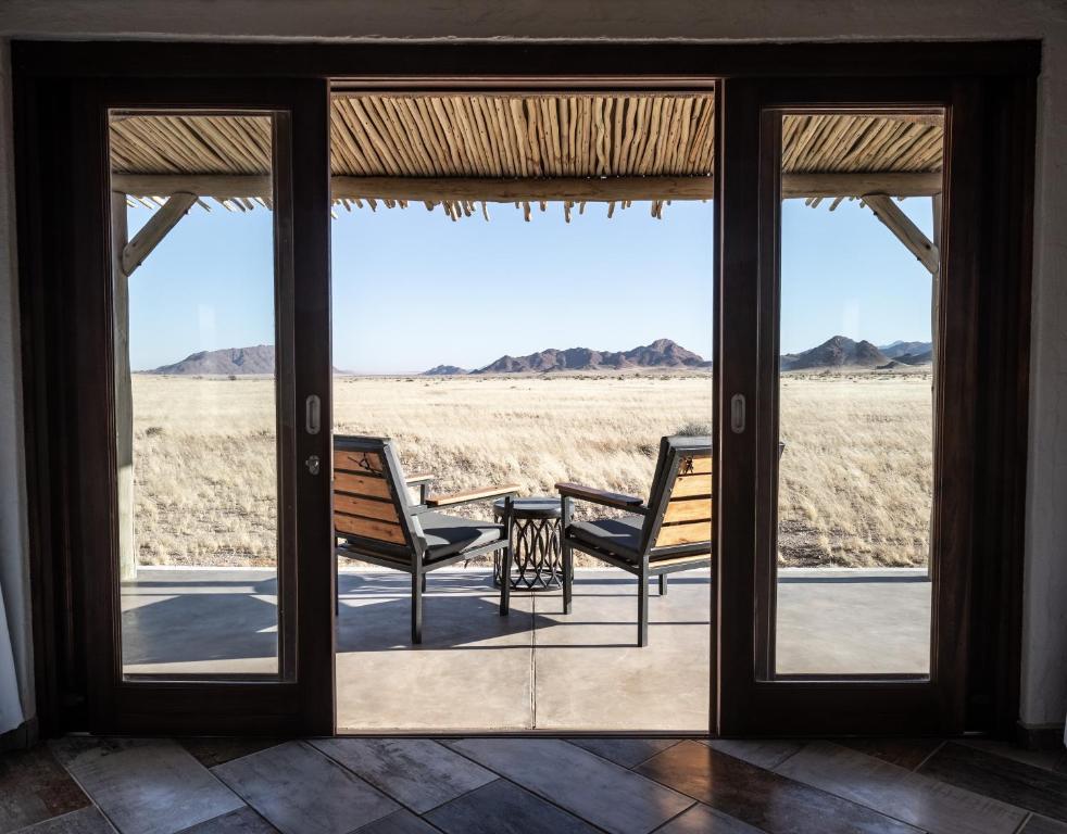 Pastoral desert view through open glass doors: two wooden chairs with a small table on a tiled patio, arid plain and distant mountains beyond.
