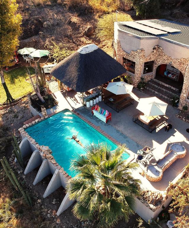 Aerial view of a resort pool area with a thatched-roof hut, umbrellas, lounge chairs, and a swimmer in a rectangular pool beside a stone villa.