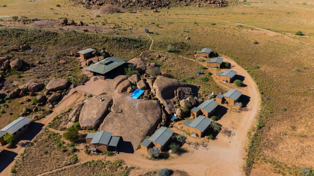 Drone view of a desert hillside camp with small brown huts and grey roofs around large boulders on a dirt road layout.