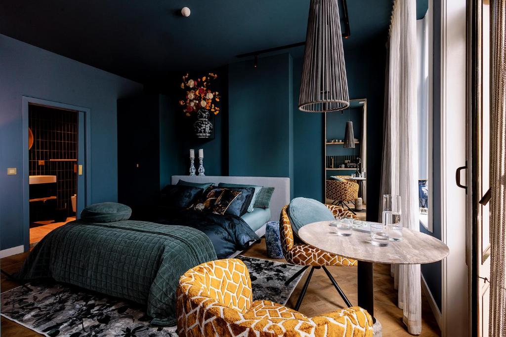 Teal-walled hotel bedroom with a king bed, green quilt, and two orange giraffe-pattern chairs around a round marble table by large windows.