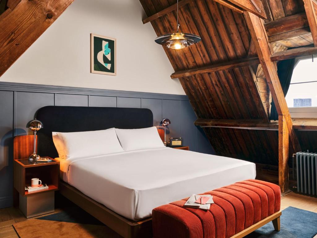 Attic bedroom with exposed wooden beams, a white bed with a navy headboard, nightstands, and a red upholstered bench at the foot.