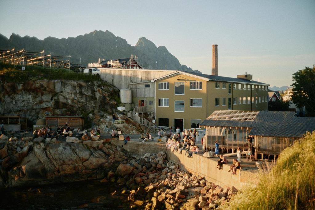 People sit and mingle on a rocky waterfront and concrete promenade beside a yellow industrial building with a tall smokestack, set against distant mountains at dusk.