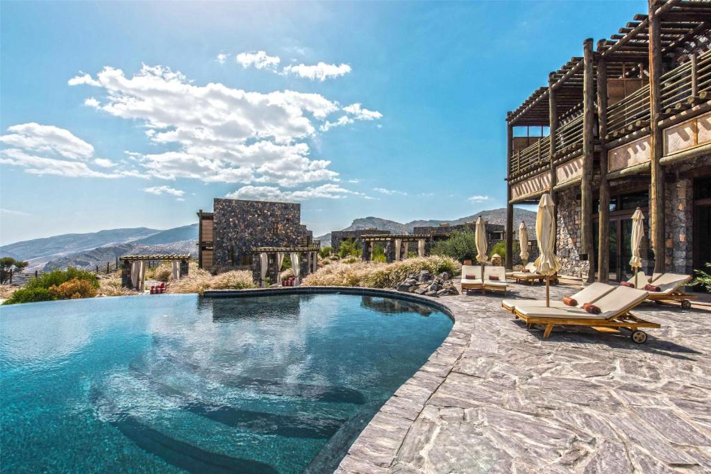Luxurious pool area at a resort with stone patio, wooden building, and sun loungers under umbrellas; mountains in the distance under a blue sky