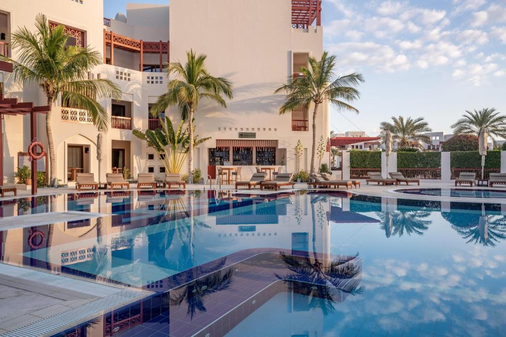Resort poolside with palm trees, white stucco buildings, and lounge chairs around a blue, reflection-filled pool.