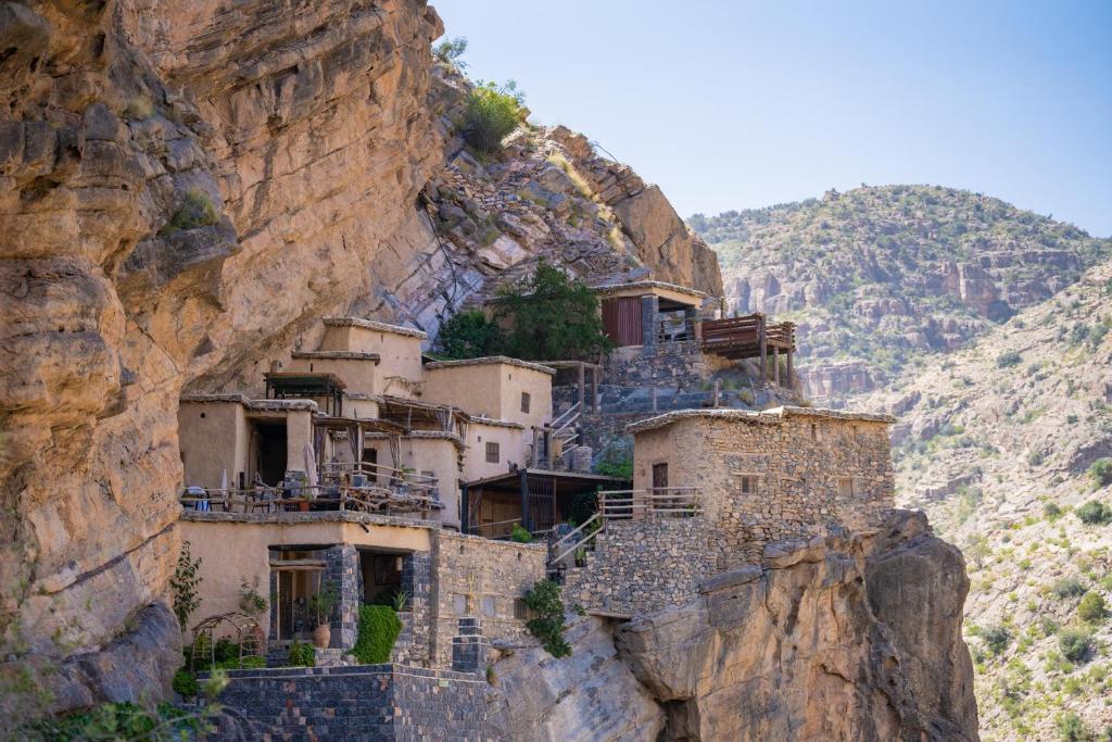 Stone houses cling to a rocky cliff, connected by stone terraces and wooden decks in a rugged mountain landscape.