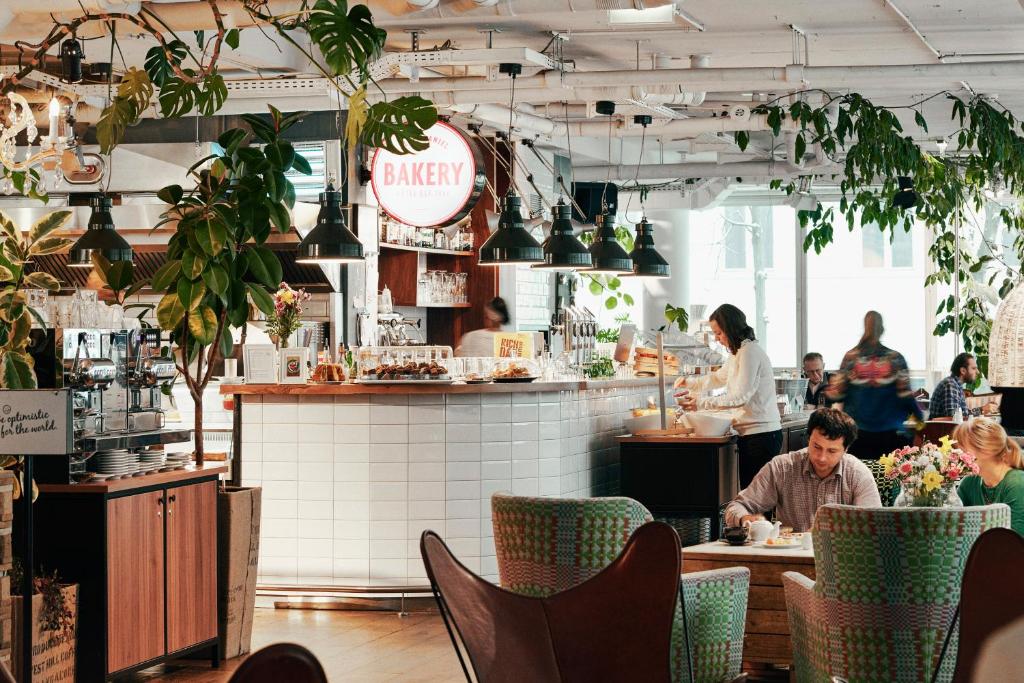 Bright bakery cafe interior with a white tiled counter, green plants, and people dining at patterned chairs in natural light.