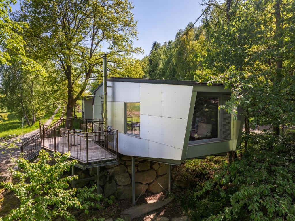Modern treehouse-like structure extends from a hillside on a metal platform with a wooden deck and railing, surrounded by green trees and clear blue sky.