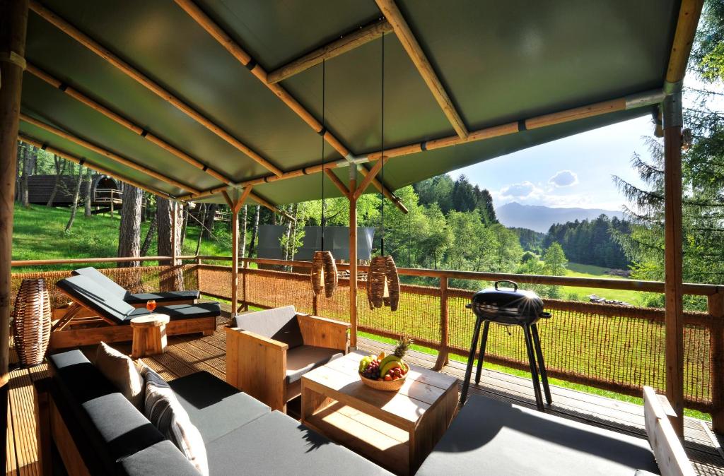 Covered outdoor deck with gray-cushioned seating, a wooden coffee table with a fruit bowl, and a kettle-style grill, overlooking a green valley.