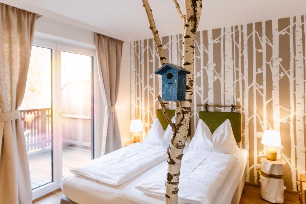 Bright hotel bedroom featuring a birch-tree motif wallpaper, a blue birdhouse on a trunk, and a neatly made bed with bedside lamps.
