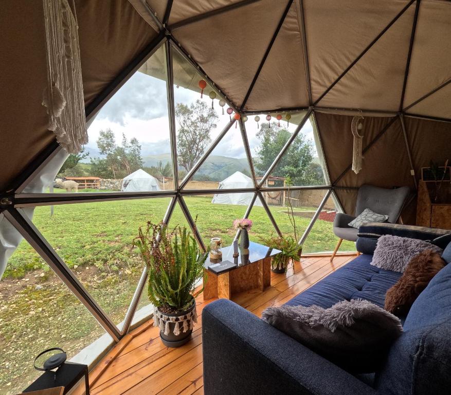 Cozy geodesic-dome living space with a blue sectional, throw pillows, potted plants, and a view of tents outside.