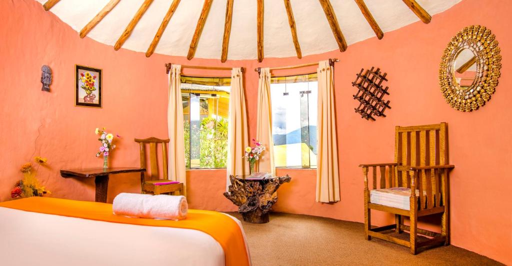 A cozy spa treatment room with coral walls, a massage table covered in white and orange linens, and a rolled towel at the end. Wooden chairs sit by windows with cream curtains, and floral arrangements add color.