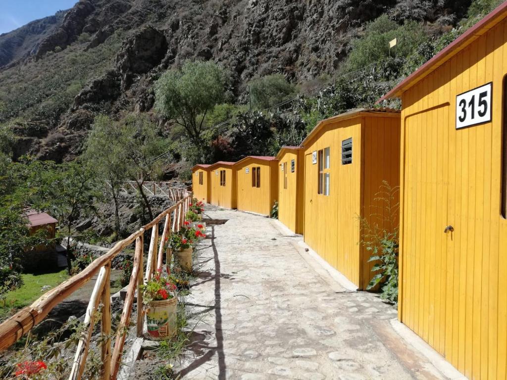 Row of yellow wooden cabins along a stone path on a mountainside, with a wooden railing and potted flowers lining the walkway.