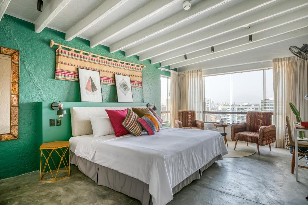 Bright hotel bedroom with a teal accent wall, white bedding, and colorful throw pillows, facing a seating area by expansive windows with a city view.