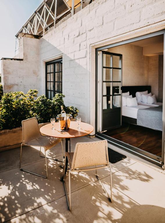 Outdoor terrace with a round table for two, a wine bucket and two glasses, wicker chairs, and open French doors to a bright bedroom.