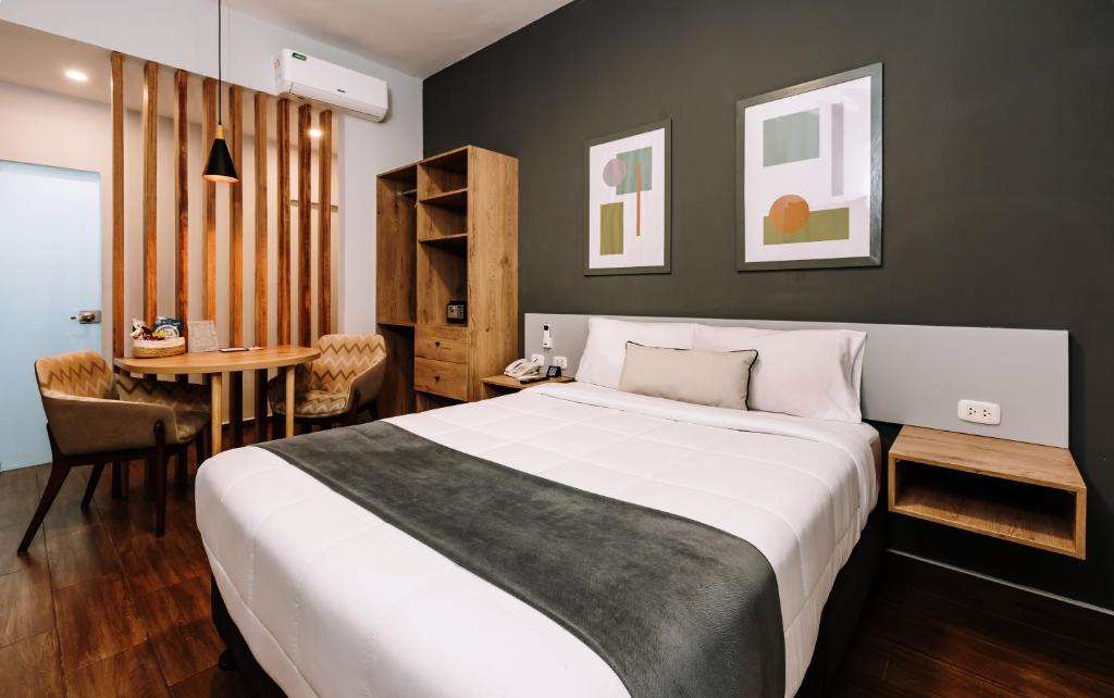Hotel room with a large white bed, gray throw, dark accent wall, and wooden nightstands; seating area with two patterned chairs nearby.