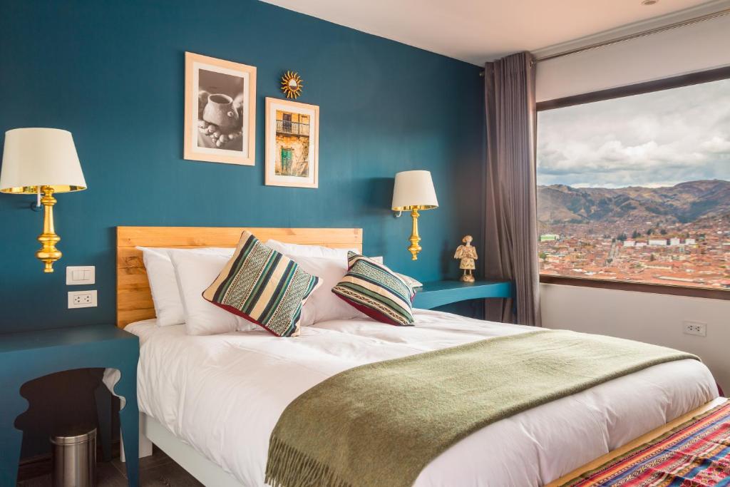Bright hotel bedroom with teal accent wall, white bed, wooden headboard, striped cushions, and gold wall lamps; large window shows city and mountains outside.