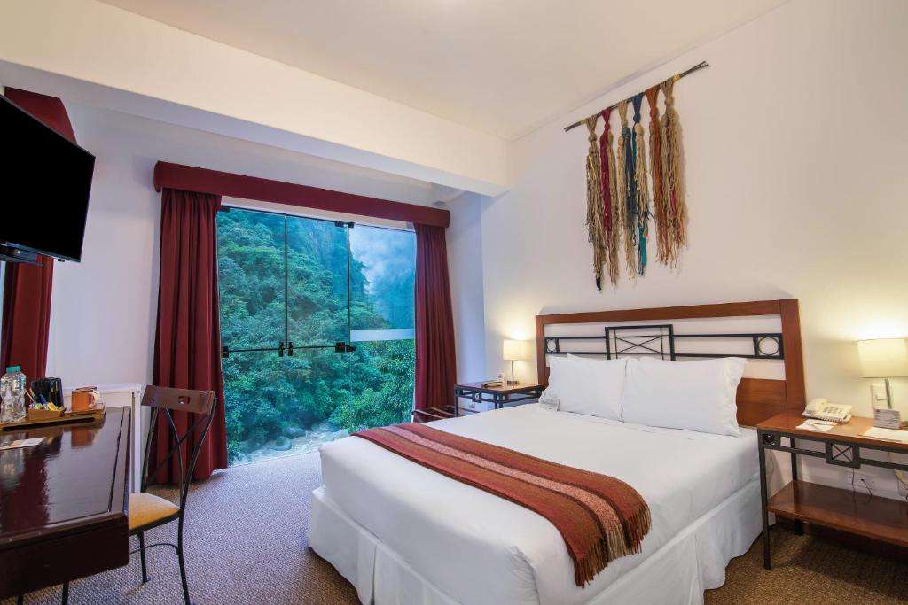 King bed in a bright hotel room with white linens, red throw, two nightstands, lamps, and a colorful wall hanging above the headboard, near a glass door to greenery.