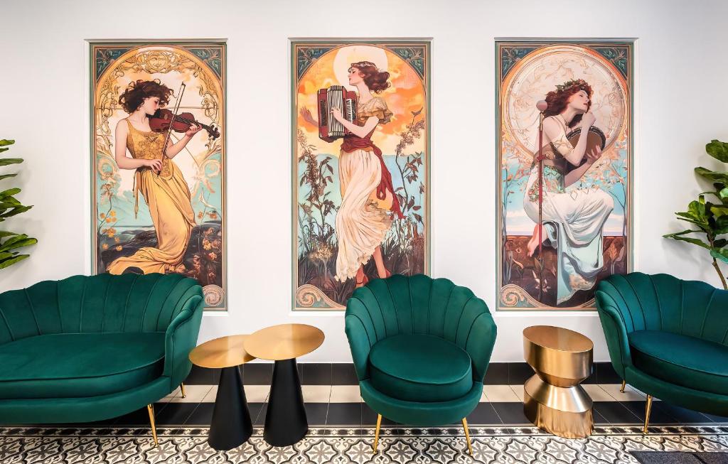 Three Art Nouveau posters of women musicians on a white wall, flanked by emerald green lounge chairs with gold tables nearby.
