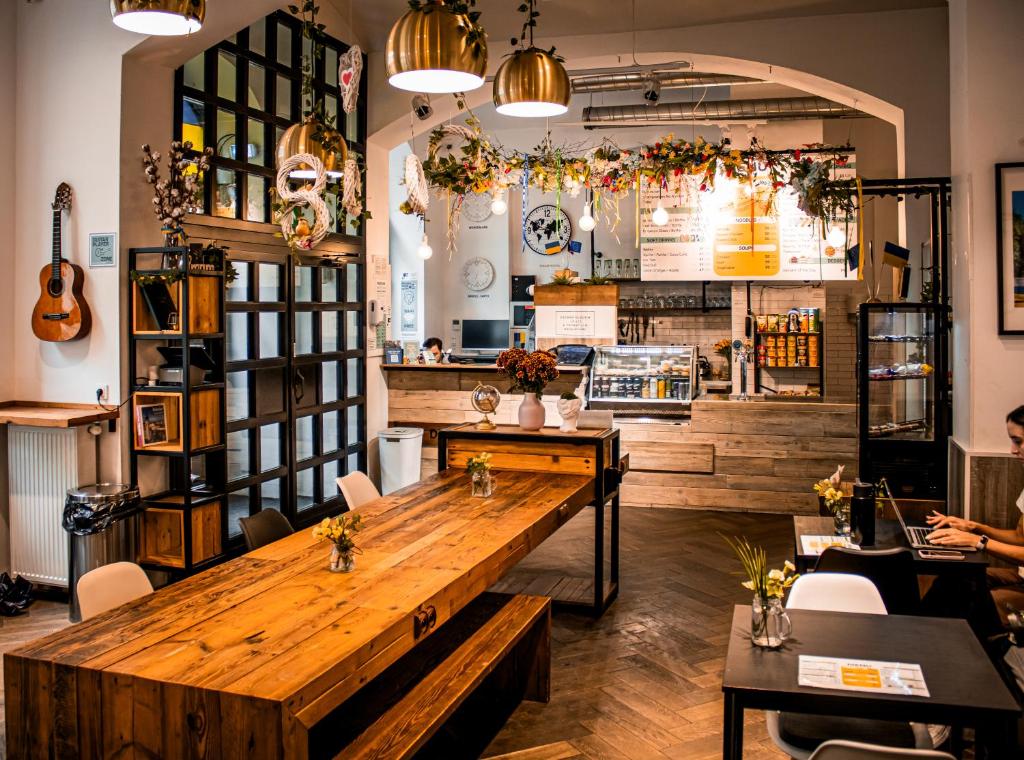 Bright cafe interior with a long wooden communal table, hanging plants, and a counter displaying pastries, under warm pendant lights.