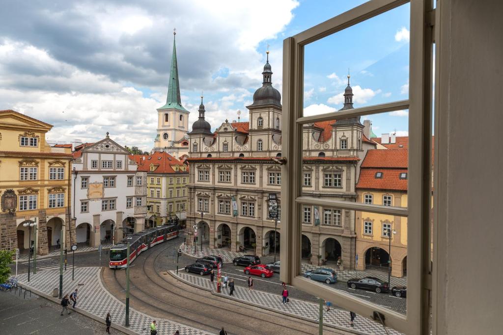 Open window frames a European city square with a red tram on curved tracks and ornate, spired buildings against a blue sky.