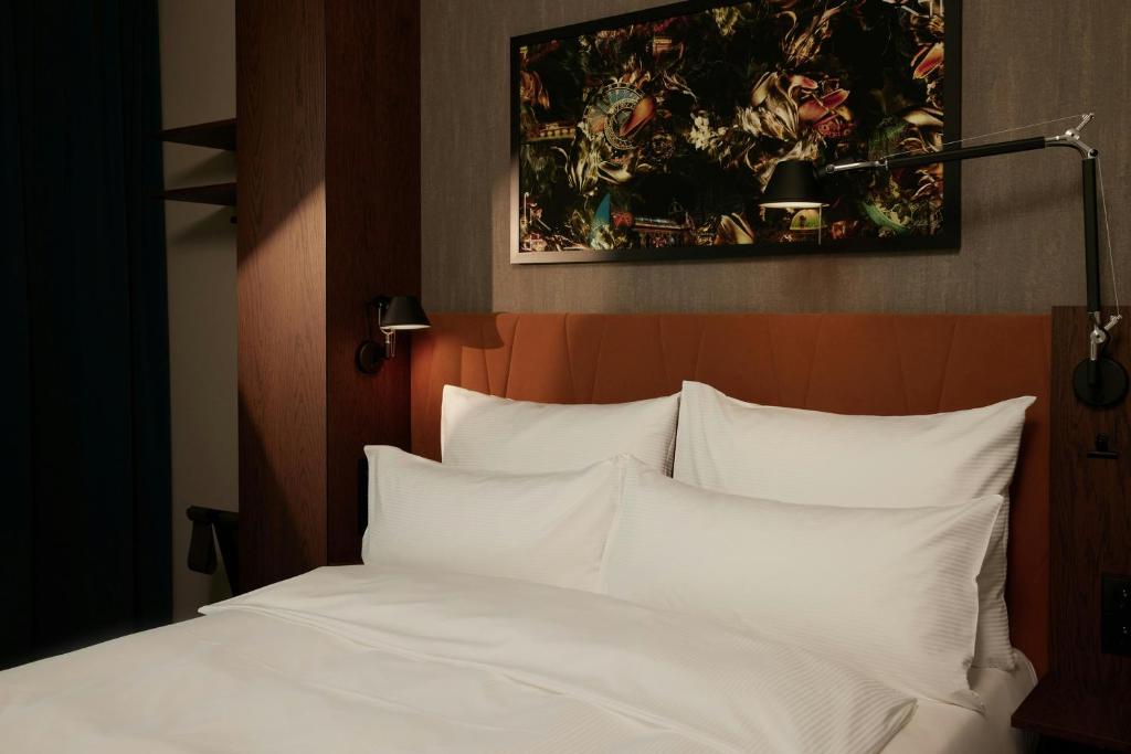 Hotel-style double bed with white linens and two large pillows against a brown upholstered headboard, wall lamps on each side, and abstract artwork above.