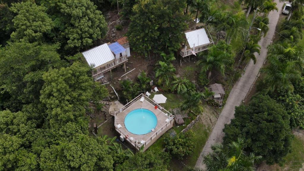 Aerial view of a circular swimming pool surrounded by a wooden deck, lounge chairs, and small cabins in a wooded area near a dirt road