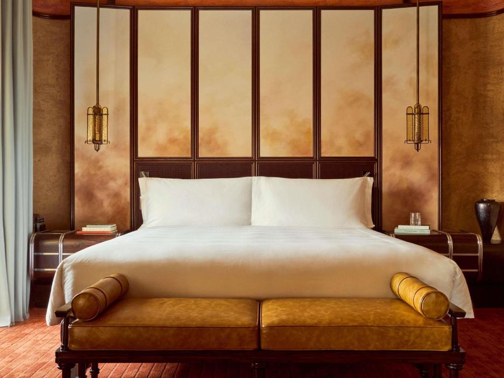 Luxurious hotel bedroom featuring a king-size bed with white linens, a tan upholstered bench at the foot, and warm amber headboard panels.