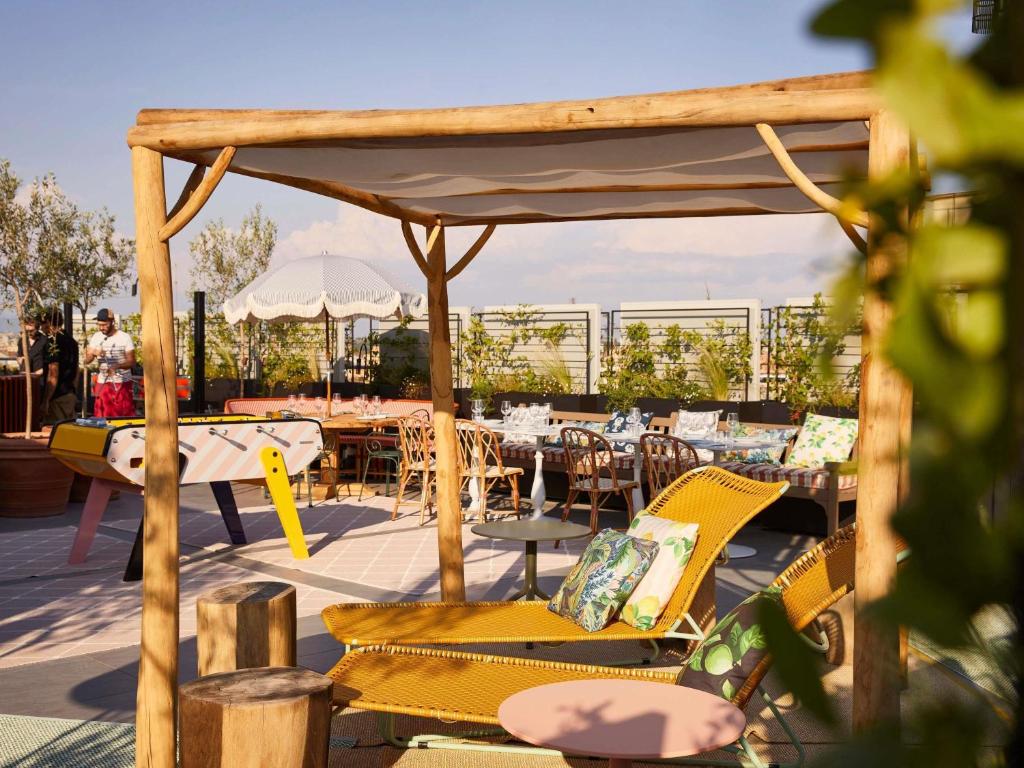 Rooftop lounge area with wooden pergola, yellow lounge chairs, and a long dining table under a canopy, sunny sky above.