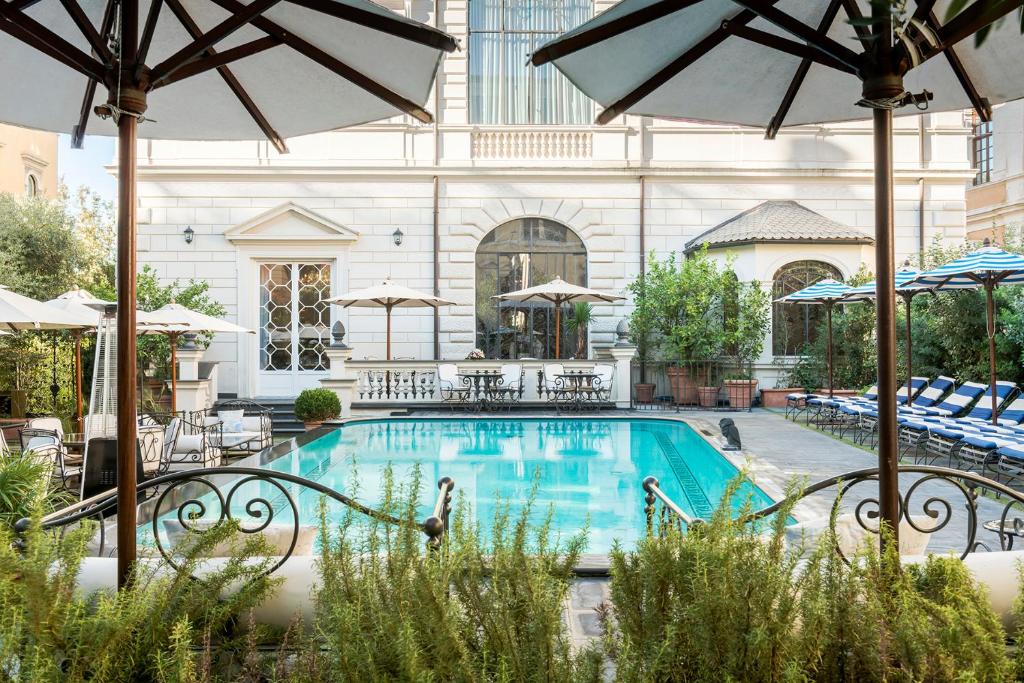 Hotel courtyard pool area with blue water, surrounded by umbrellas and lounge chairs against a pale stone building.