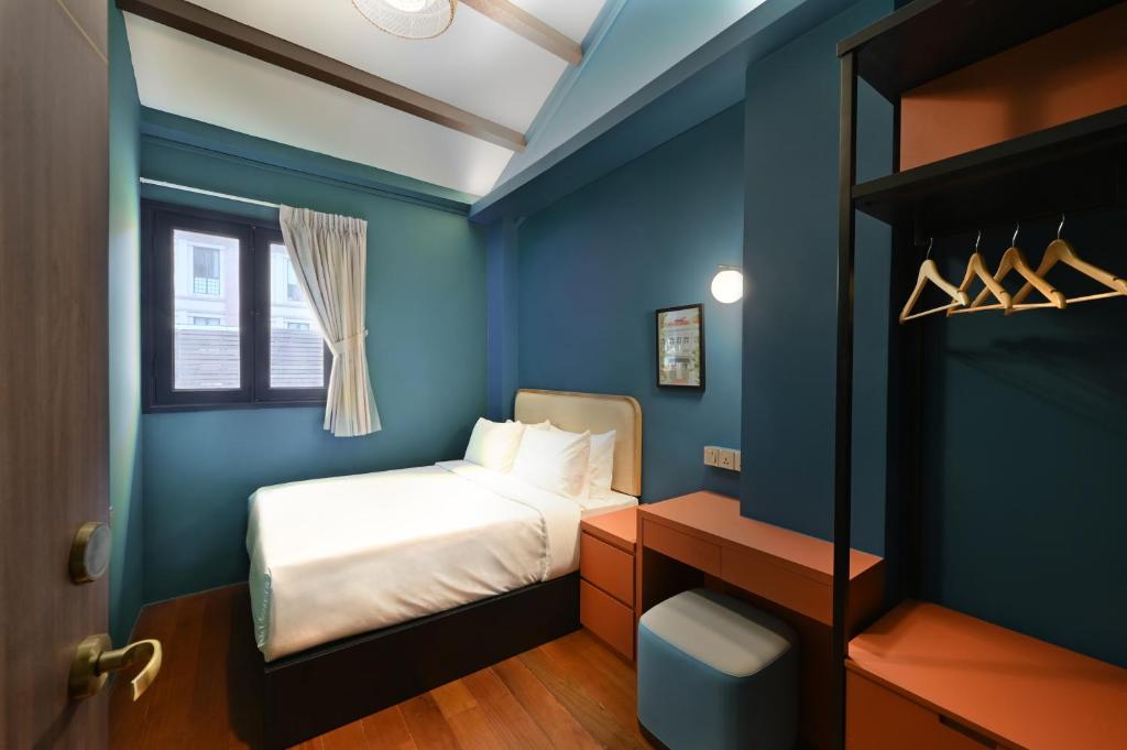 Small blue hotel room with a white-bedding bed, window with curtains, desk, and an open wardrobe with hangers.