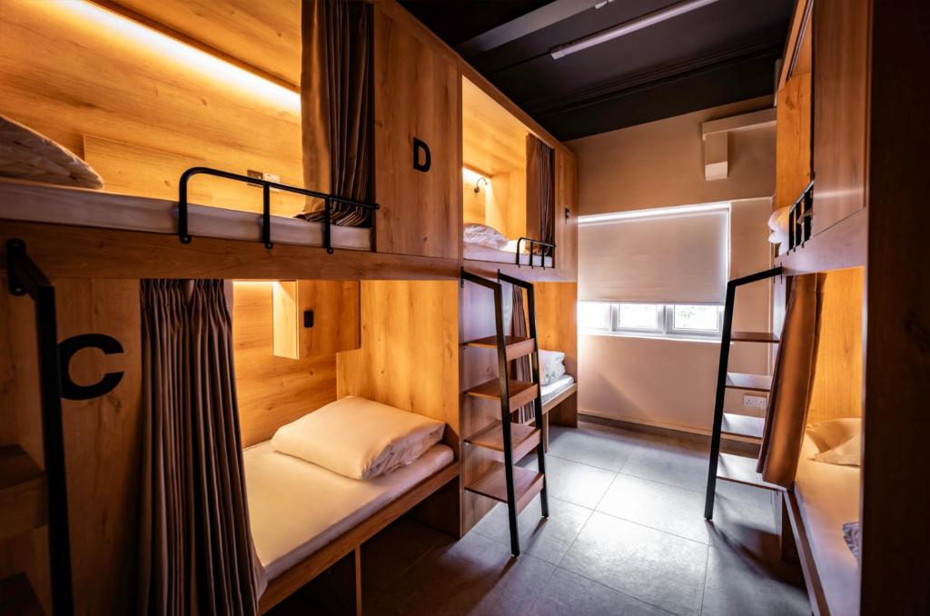 Wooden hostel dorm with four bunk beds, privacy curtains, and labeled cubbies along a warm-lit wall. A window sits at the far end.
