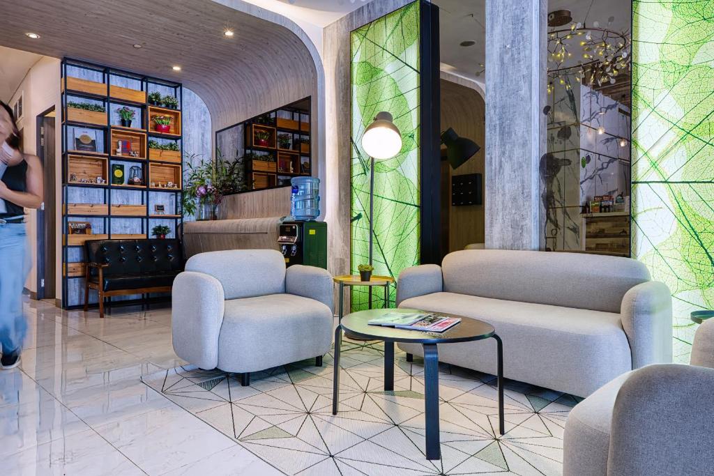 Modern hotel lobby lounge with gray sofas, a round coffee table, and a geometric tiled floor. Exposed shelving and a water cooler back left add to the space.