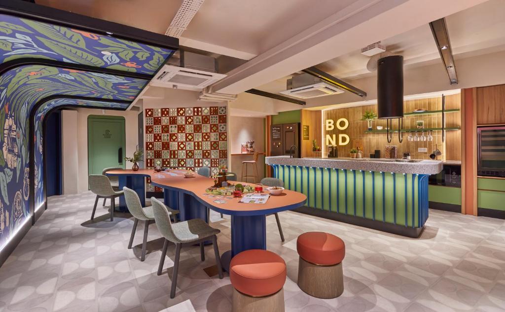 Colorful, modern lounge with a long communal table, grey chairs, and a blue-green striped bar next to a patterned wall mural on the left.