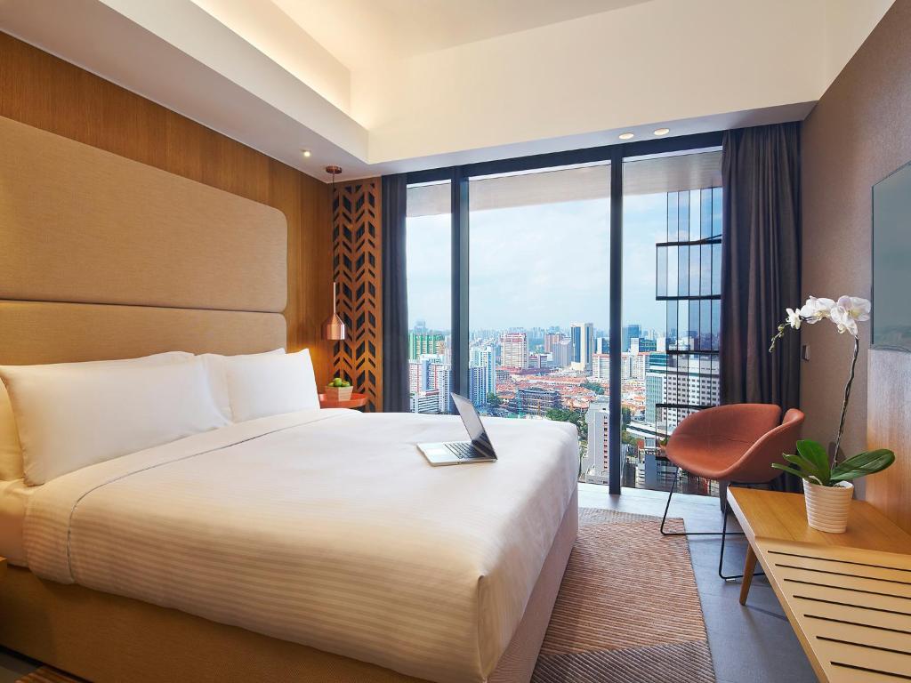 Hotel room with a large bed, bedside lamps, and a laptop on the duvet, overlooking a city skyline through floor-to-ceiling windows.