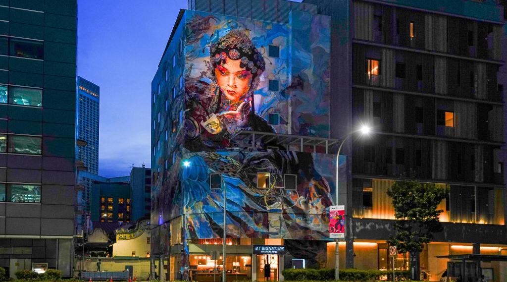 Nighttime street view of a building with a large mural of a woman's face on its facade, glowing blue and purple hues.
