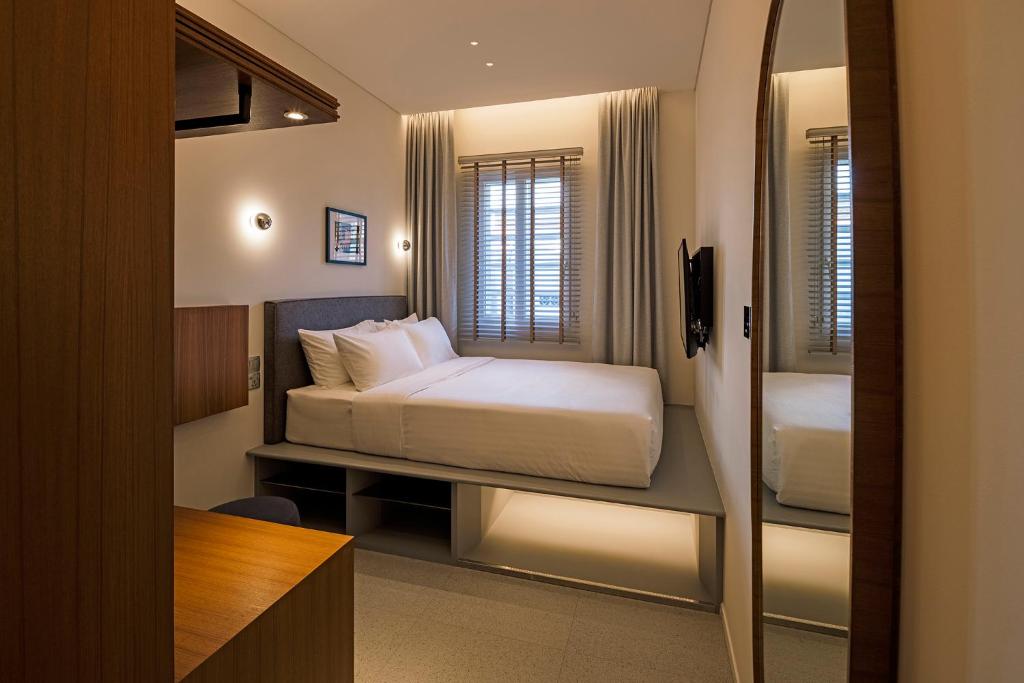 Hotel room with a white bed on a raised platform, window with blinds, wall-mounted TV, and a wooden desk nearby.